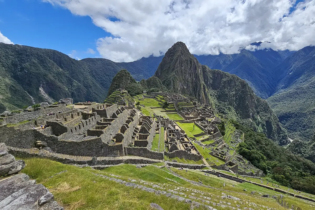 The stone terraces and buildings of Machu Picchu on a green ridge, with the steep peak of Huayna Picchu rising behind under broken cloud